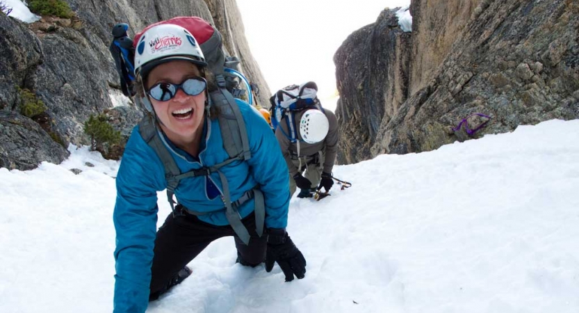 a person wearing mountaineering gear looks up and smiles at the camera while making their way up a snowy incline between two large rocks. There is another person in mountaineering gear behind them.